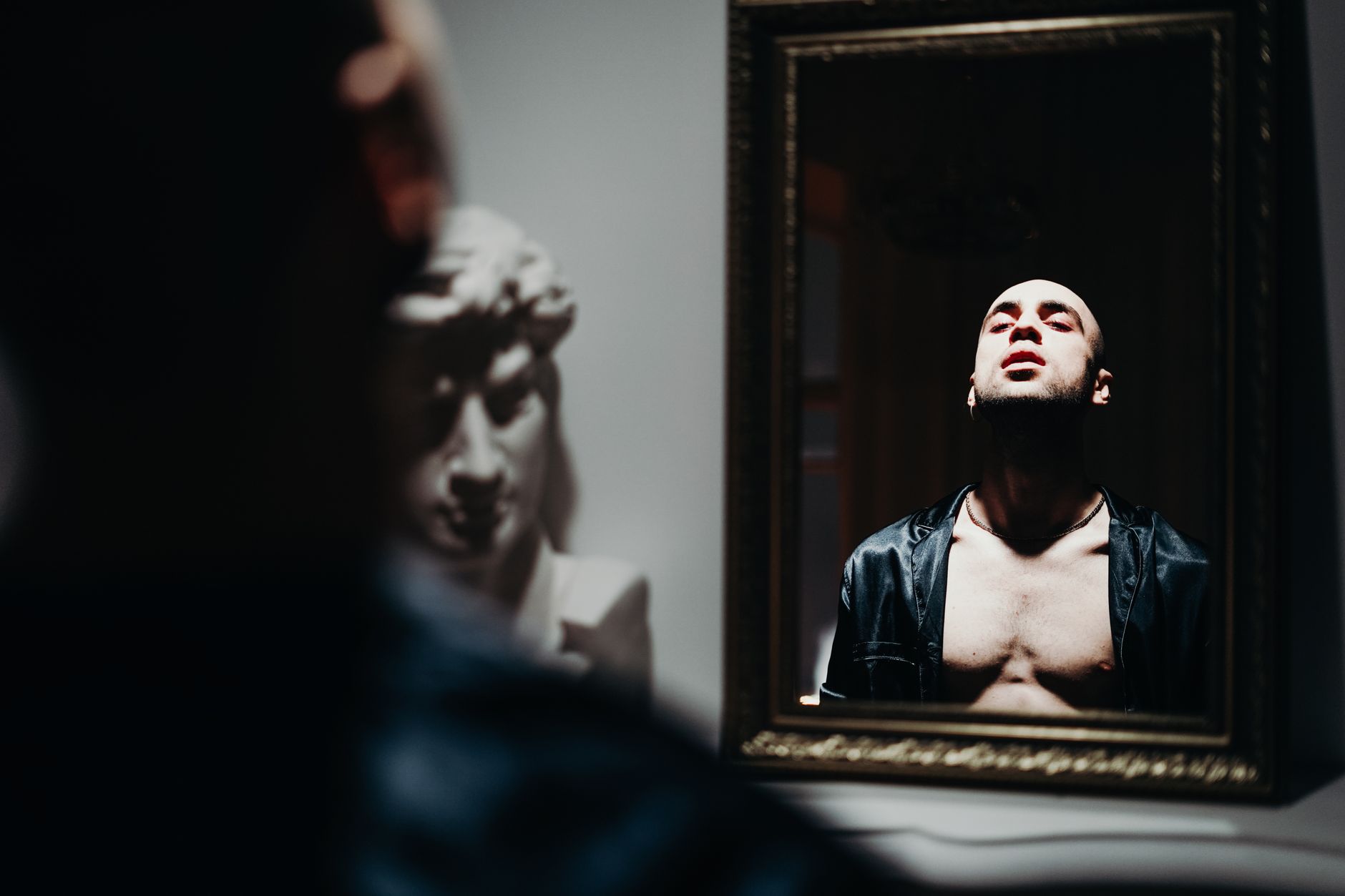 A person gazes at their reflection in a mirror, their face partially obscured by shadow and darkness.